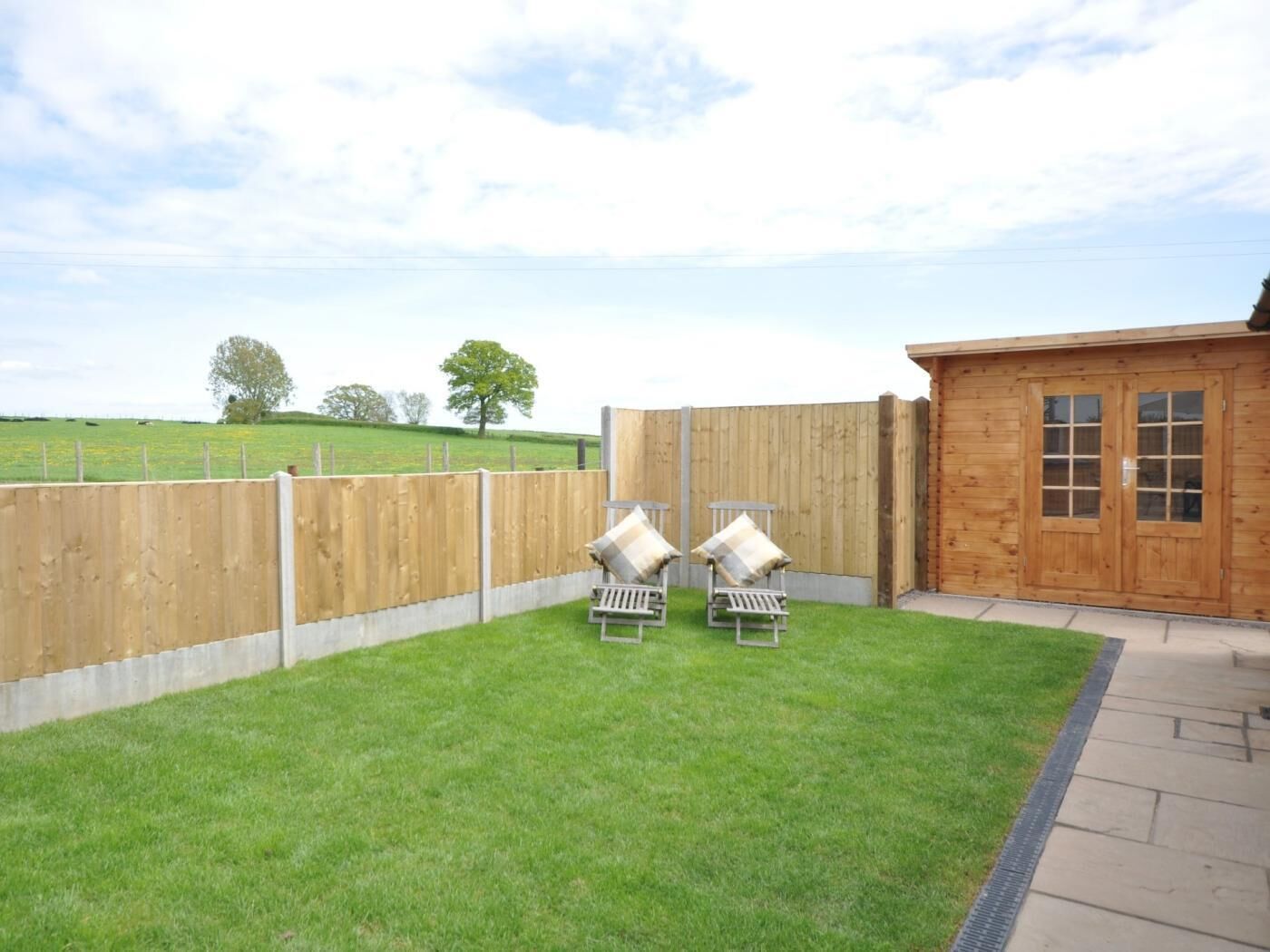 A wooden building with a door and windows, a fenced yard with two lounge chairs, and a grassy field in the background.