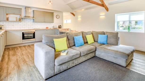 A modern kitchen with a built-in oven, a grey sectional sofa with colorful cushions, and a window with a view of greenery. - 2 Bed in Barnstaple (oc-60443) (Bittadon)
