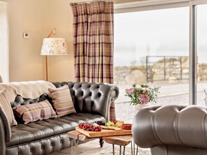 A leather sofa with a blanket and pillows, a wooden side table with a fruit platter, and a view of the outdoors through a window with curtains. - 4 Bed in Betws-y-coed (oc-60172) (Pentrefoelas)
