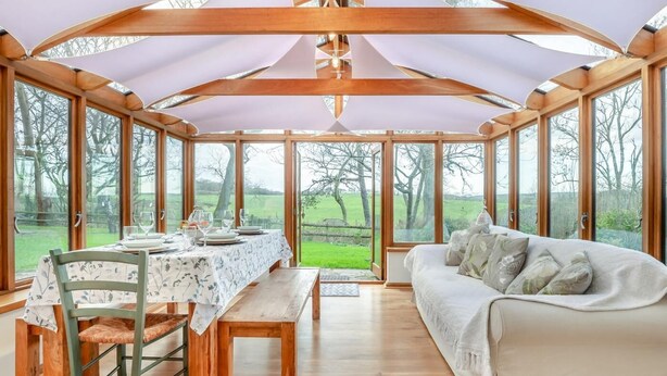 Uma estufa com uma mesa de jantar posta, um sofá e grandes janelas com vista para o exterior.