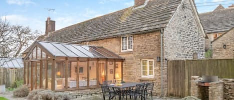 A stone house with a glass conservatory, a patio with a table and chairs, and a wooden fence.