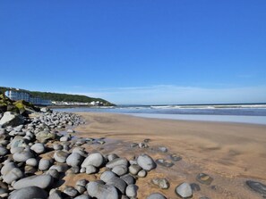 Une plage bordée d’une côte rocheuse, un immeuble à plusieurs étages et un ciel bleu dégagé.