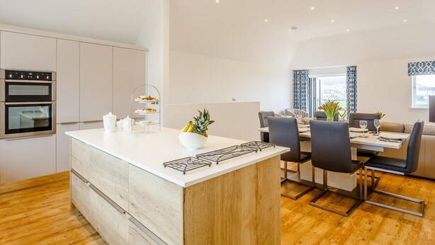 A modern kitchen with built-in ovens, a central island with a gas stove, and a dining area with a table and chairs. - 3 Bed in Westward Ho! (oc-60678) (Westward Ho!)
