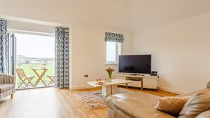 A living room with a sofa, a TV, a dining table with chairs, and a view through a glass door to an outdoor area. - 3 Bed in Westward Ho! (oc-60678) (Westward Ho!)