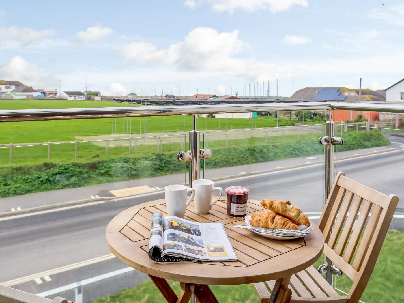 Uma mesa de madeira redonda com um jornal, duas canecas brancas, um pote de geleia e um prato de croissants numa varanda com vista para uma rua arborizada e edifícios ao longe.