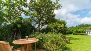 Eine Holzterrasse mit einem runden Tisch und Stühlen, umgeben von üppiger Bepflanzung und einem weißen Pavillon im Hintergrund.