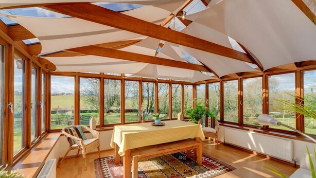 A conservatory with wooden beams, large windows, a dining table, chairs, and a patterned rug. - 3 Bed in Corfe Castle (oc-ic001) (Corfe Castle)