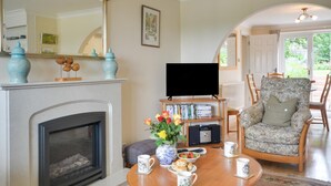 Una sala de estar con chimenea, televisor, sillón floral y una mesa de centro con un jarrón de flores.