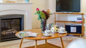 Una sala de estar con chimenea, televisor, una mesita con revistas, un jarrón con flores y un juego de té.