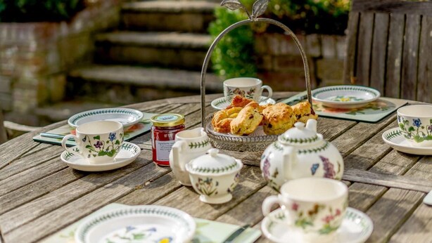 Una mesa de madera dispuesta con tazas de té, platitos, una tetera, un azucarero y una cesta de scones.