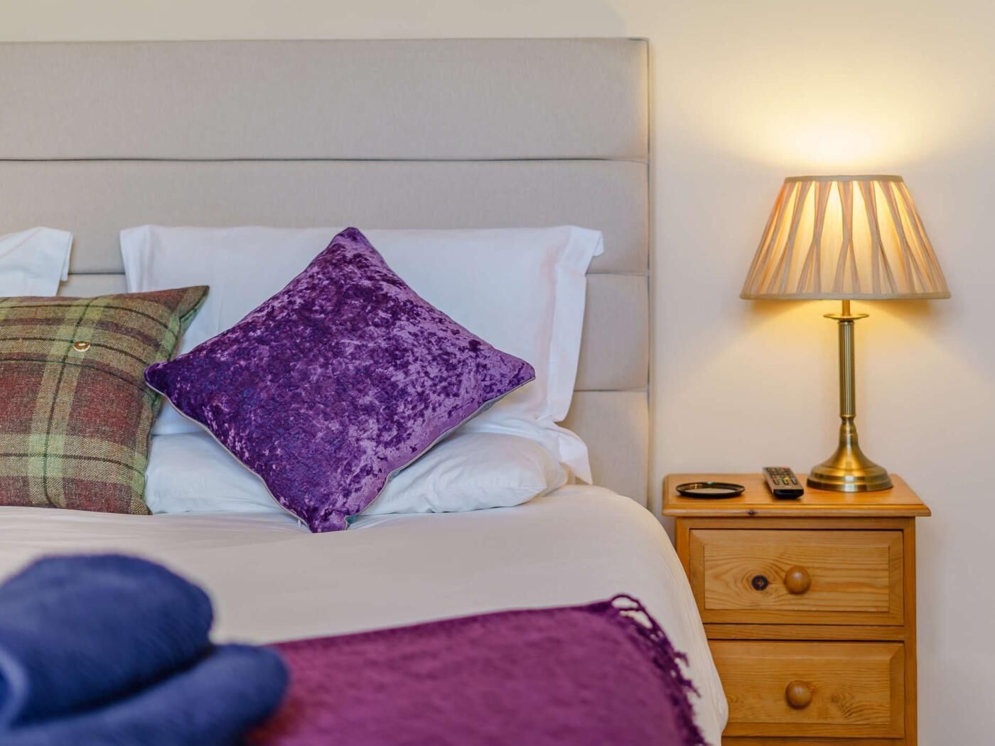 Een opgemaakt bed met hoofdbord, een paars kussen, een ruitdessin kussen, een houten nachtkastje met lamp en een afstandsbediening.