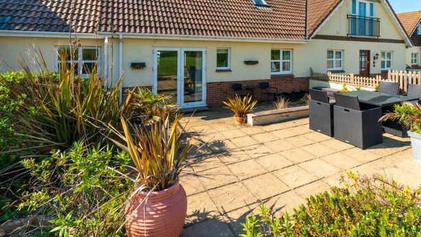 Ein zweistöckiges Haus mit einem Ziegeldach, einer Terrasse mit Gartenmöbeln und einem Garten mit verschiedenen Pflanzen.