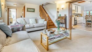A living room with a sofa, a coffee table with newspapers, a staircase, and a kitchen area. - 3 Bed in Carisbrooke (oc-ic037) (Newport)