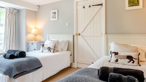 Una habitación de hotel con dos camas, una puerta blanca grande, una mesita de noche con una lámpara y un cuadro en la pared.