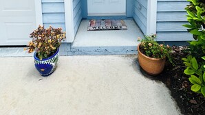 Une maison bleue avec une porte blanche, le numéro « 66 » sur la porte, un paillasson et deux plantes en pot.
