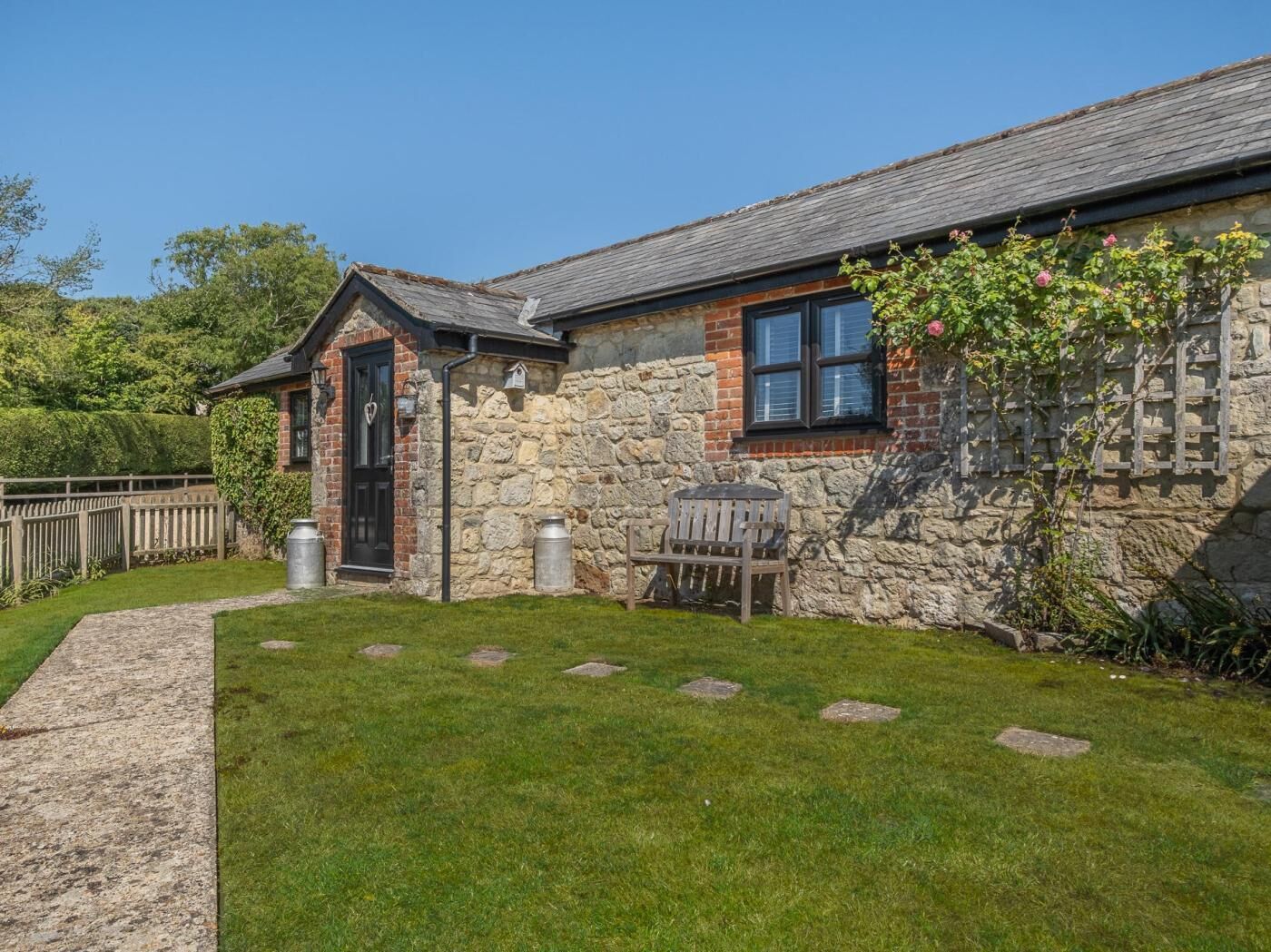 A stone cottage with a small garden, a wooden bench, and a metal barrel.
