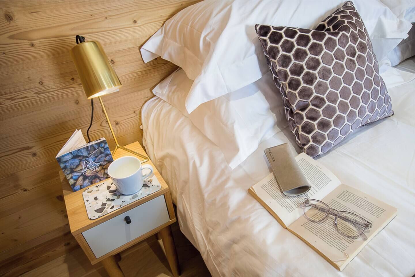 A neatly made bed with a white comforter, a patterned pillow, a bedside lamp, a book, and a cup on a wooden nightstand.