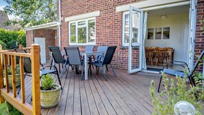 An outdoor patio with a wooden deck, a table with chairs, and a brick building with a glass door. - 4 Bed in Shanklin (oc-ic068) (Shanklin)