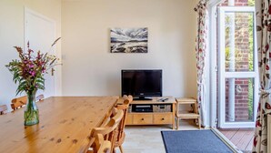 A dining area with a wooden table and chairs, a television, and a window with curtains. - 4 Bed in Shanklin (oc-ic068) (Shanklin)