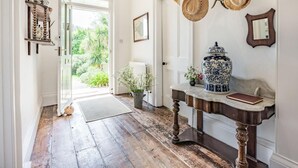 Een hal met een houten vloer, een witte deur die naar buiten leidt, een tafel met een vaas en een blauwe en witte keramieken pot.