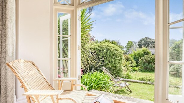 Een kamer met rieten meubels, een glazen deur die uitkomt op een tuin en uitzicht op groen.