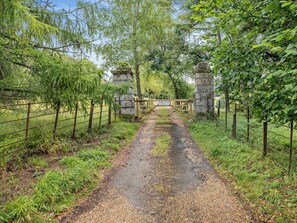 Camino rural que lleva a una puerta de piedra con un cartel, rodeado de follaje y cercas.
