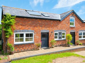 A brick building with a sloped roof, multiple windows, and a small garden area with potted plants. - 2 Bed in Alverstone (oc-ic091) (Alverstone Garden Village)