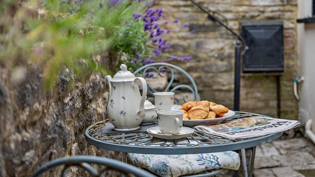 Una mesa con tetera, tazas y croissants, frente a una pared de piedra con flores moradas.