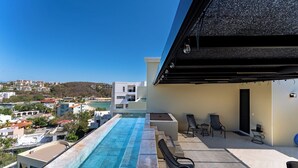 Ein Pool auf dem Dach mit Blick auf die Stadt, eine überdachte Terrasse mit Sitzgelegenheiten und ein modernes Gebäude im Hintergrund.