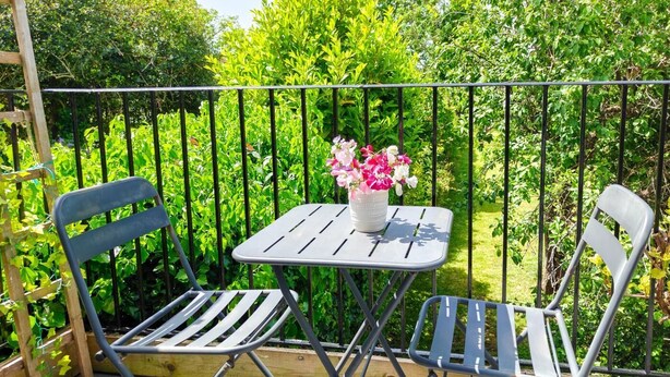 Ein Balkon mit zwei Metallstühlen und einem kleinen Tisch, umgeben von einem Metallgeländer und üppigem Grün.