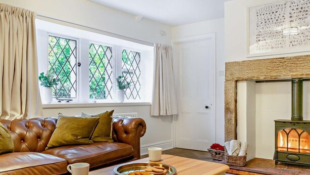 A cozy living room with a leather sofa, a wooden coffee table with mugs and cookies, a fireplace, and a window with a lattice design. - 2 Bed in Alnwick (oc-93093) (Lemmington Hall)