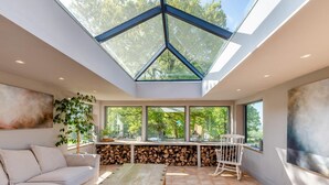 Un interior moderno con techo alto, ventanas amplias, un sofá, una mesa de madera y una pila de leña.