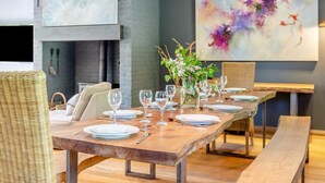 Comedor con una larga mesa de madera dispuesta para seis personas, flanqueada por sillas de mimbre, un banco y una chimenea.