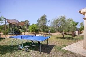 A blue outdoor ping pong table with a canopy and a trampoline in a grassy area with trees.