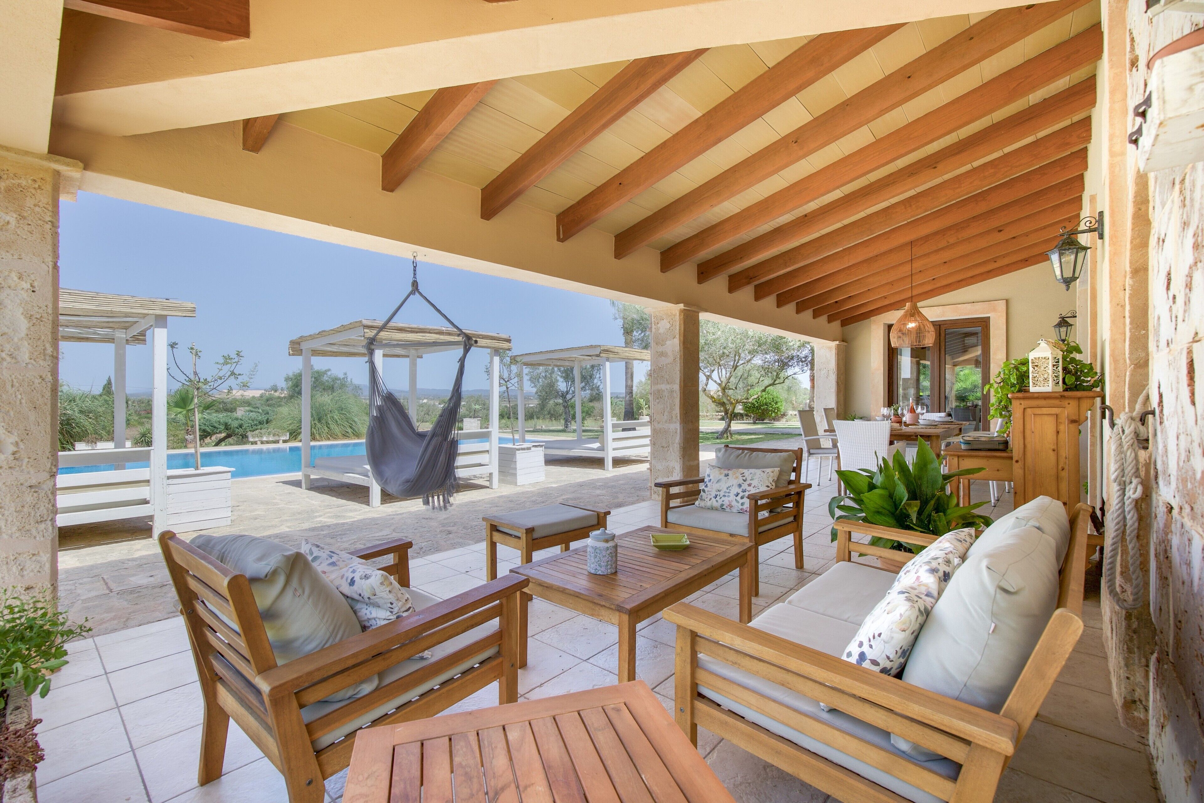 Un espacio de estar al aire libre techado con muebles de madera, una hamaca colgante y vistas a una piscina.