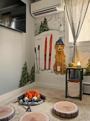 A room with a ski-themed wall mural, a small table with a camera and candle, and a dog bed with a plush toy. - GameRoom•FirePit•Grill•KidsFriendly•9 min Kalahari (Long Pond)