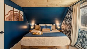 Quarto com teto de madeira, cama com colcha acolchoada, almofadas, mesa de cabeceira com abajur e janela com vista para árvores.