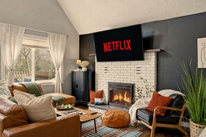 Un salon avec une cheminée, un téléviseur fixé au mur et une fenêtre avec des rideaux.
