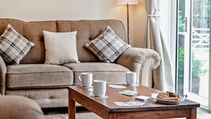 A beige sofa with plaid cushions, a wooden coffee table with mugs and playing cards, and a lamp in a room with a large window. - 3 Bed in Sturminster Newton (oc-rcorn) (Sturminster Newton)
