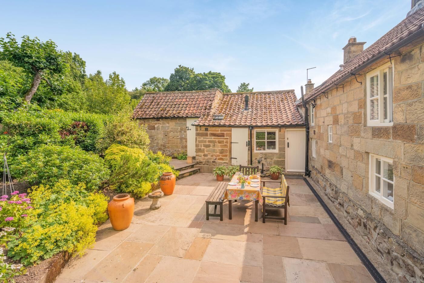 A stone building with a tiled roof, a patio with a table and chairs, and potted plants.
