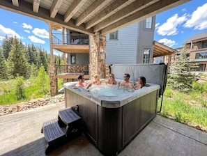 A hot tub with six people, a stone pillar, and a wooden deck. - The Riverstone | Luxury Alcove • Kindred • Ski-In/Out • Spa • Heated Garage (Keystone)
