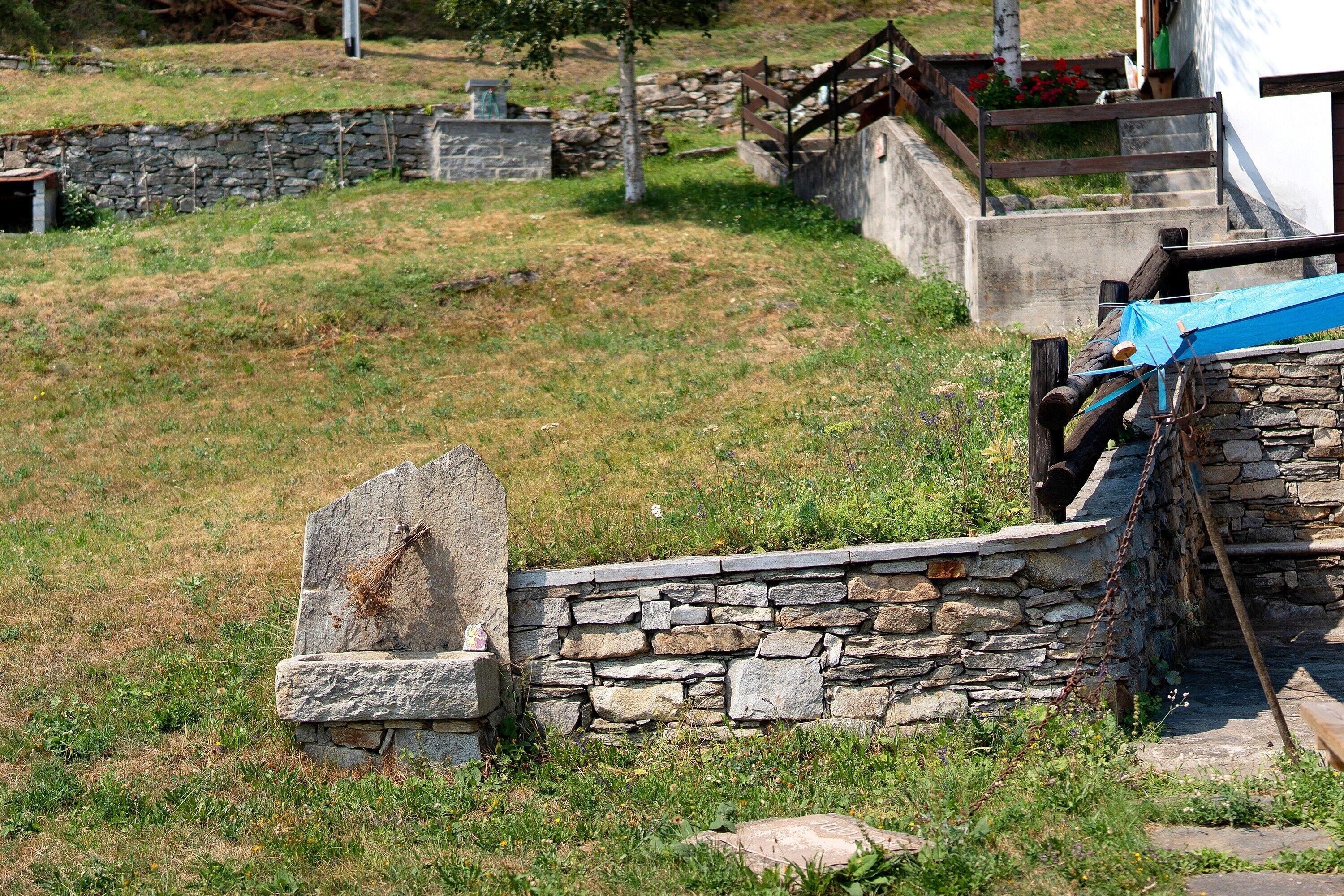A stone wall with a gravestone, a wooden bench, and a metal structure in a grassy area.
