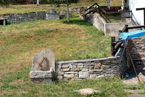A stone wall with a gravestone, a wooden bench, and a metal structure in a grassy area. - Mountain Hut 'la Fraccia' in pristine Alpine setting (Trasquera)