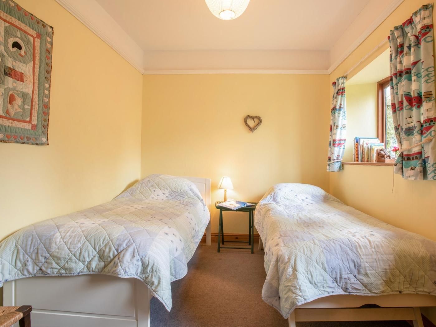 A room with two single beds, a small table with a lamp, a heart-shaped decoration on the wall, and a window with curtains.