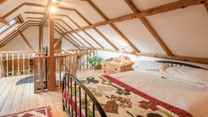 Ein Schlafzimmer unter der Dachschräge mit hohen Decken, freiliegenden Holzbalken, einer Holztreppe, einem Bett mit Quilt und einem kleinen Tisch mit Lampe.