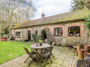 A rustic stone house with a tiled roof, a brick chimney, and a round wooden table with chairs in the yard. - 3 Bed in South Creake (oc-kt099) (South Creake)