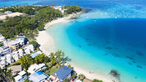 Vista aérea de una zona costera con playa, aguas azules cristalinas y un pequeño grupo de edificios.