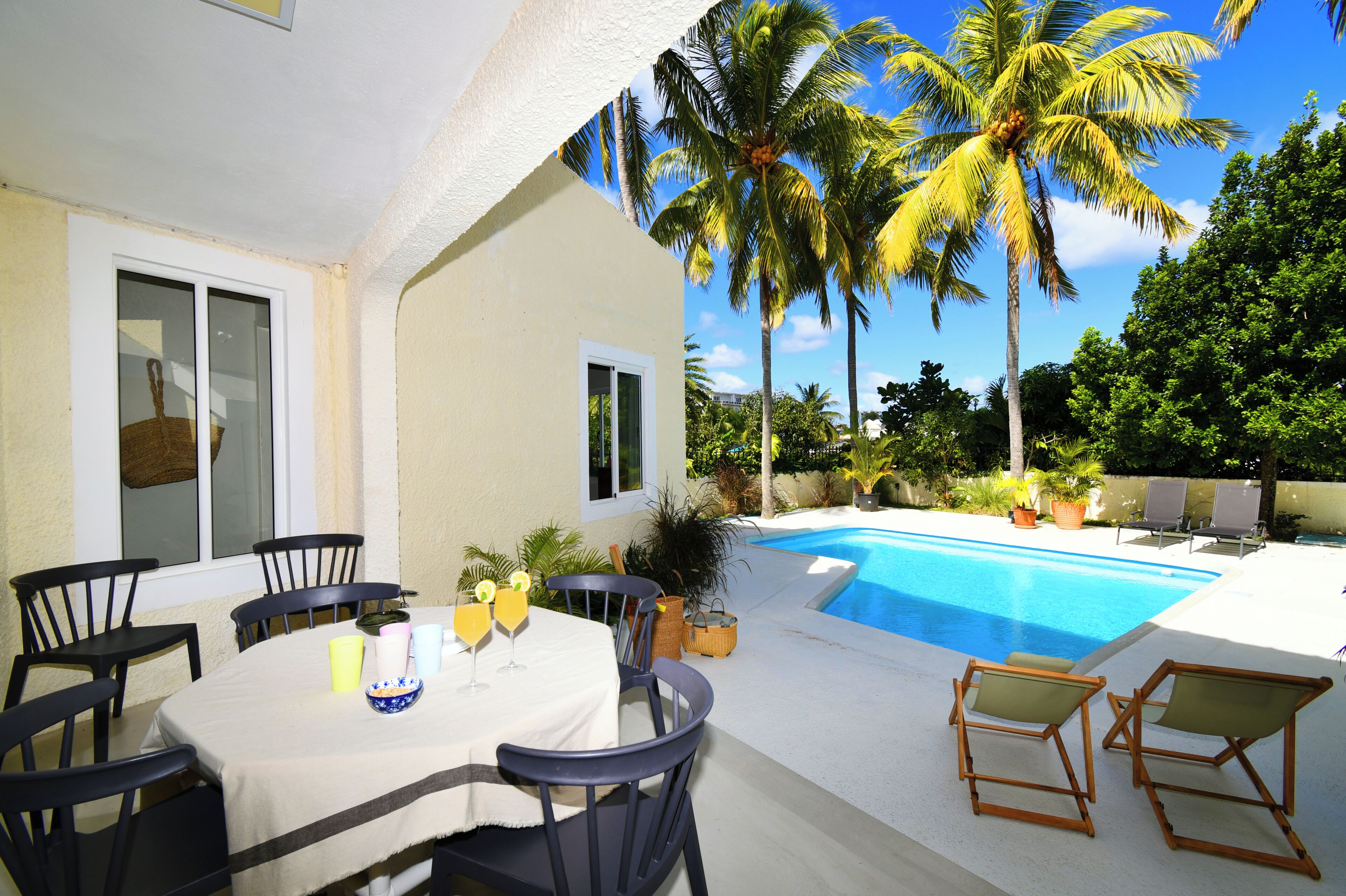 An outdoor pool area with chairs and a table, surrounded by palm trees and greenery.