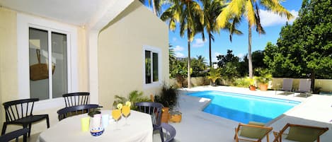 An outdoor pool area with chairs and a table, surrounded by palm trees and greenery.