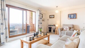 A living room with a sofa set, a coffee table, a dining table with a vase of flowers, and a view of a brick wall through a sliding door. - 2 Bed in Bideford (oc-oce16) (Westward Ho!)